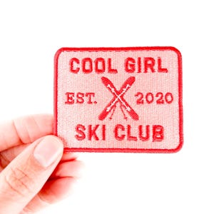 Puede incluir: Un parche bordado rojo con el texto "COOL GIRL SKI CLUB EST. 2020" y esquís cruzados. El parche es cuadrado con esquinas redondeadas y se sostiene sobre un fondo blanco.