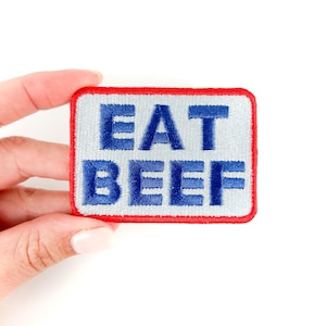 May include: A white iron-on patch with a red border. The patch has blue embroidered text that reads "EAT BEEF".