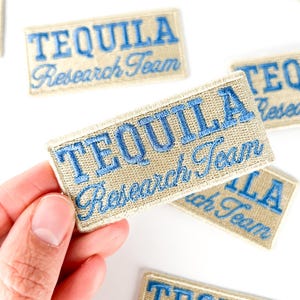 Può includere: Toppa rettangolare ricamata con le parole "TEQUILA Research Team" in filo blu su sfondo beige. Diverse toppe sono visibili, una tenuta in mano.