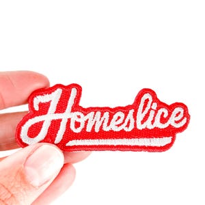 May include: A red embroidered patch with the word "Homeslice" in white script. The patch has a white border and is held by a hand against a white background. The patch is a fun accessory.