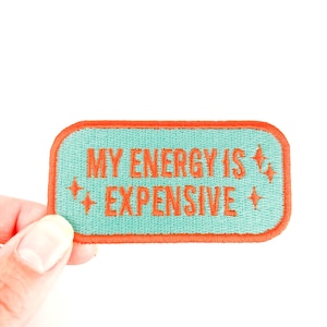 Op de afbeelding: Een rechthoekige patch met de tekst "MY ENERGY IS EXPENSIVE" in oranje letters. De patch heeft een turquoise achtergrond met een oranje rand en sterontwerpen.