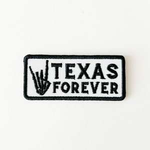 Puede incluir: Parche blanco bordado con puntadas negras. El parche presenta un gesto de mano con el índice y el meñique extendidos y los demás dedos cerrados, junto con el texto "TEXAS FOREVER".