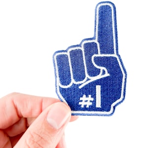 May include: A blue foam finger with a white outline and the number 1. The foam finger is held up, showing the index finger. The background is white.