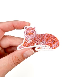 May include: Embroidered patch of a tiger in orange and pink. The tiger is in a reclining pose with detailed stitching. The patch has a white border. The patch is held against a white background.