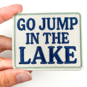 Puede incluir: Parche cuadrado bordado con el texto "GO JUMP IN THE LAKE" en hilo azul marino sobre un fondo blanquecino. El parche tiene un borde verde claro y está sujeto por una mano.