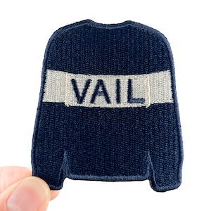 Op de afbeelding: Een marineblauwe geborduurde patch in de vorm van een trui. Het woord "VAIL" is wit in het midden geborduurd. De patch wordt door een hand tegen een witte achtergrond gehouden.