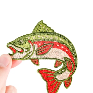 May include: Embroidered patch of a leaping fish, featuring a vibrant color scheme. The fish has a green back with white spots, a red stripe along its side, and red fins and tail. The patch is held against a white background.