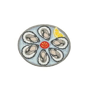 May include: A circular embroidered patch featuring six grey and white oysters arranged in a flower-like pattern on a light blue background. A small yellow lemon wedge is positioned at the top right, and a red circular garnish sits in the center.