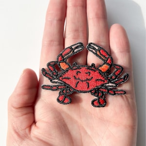 Op de afbeelding: Rode geborduurde krabbenpatch met zwarte contouren. De krab heeft een glimlachend gezicht en staat in een looppositie.