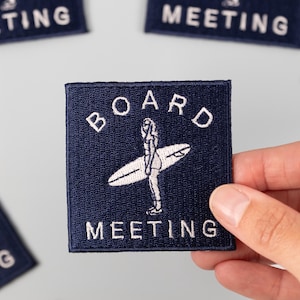 Op de afbeelding: Marineblauw geborduurd patch met de woorden "BOARD MEETING" en een surferafbeelding. De patch is vierkant en wordt door een hand vastgehouden. Andere patches met het woord "MEETING" staan op de achtergrond.