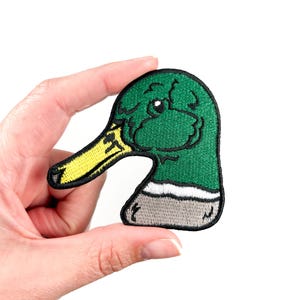 Puede incluir: Parche bordado verde y amarillo de la cabeza de un pato con una franja blanca alrededor del cuello.