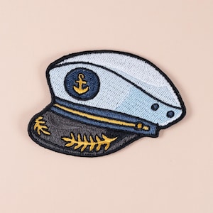 Könnte beinhalten: Gestickter Aufnäher eines Kapitäns-Hutes. Der Hut hat eine hellblaue Krone, einen dunkelblauen Rand mit goldenen Blattdetails und ein goldenes Ankeremblem. Der Aufnäher hat einen schwarzen Rand und ist vor einem hellrosa Hintergrund platziert.