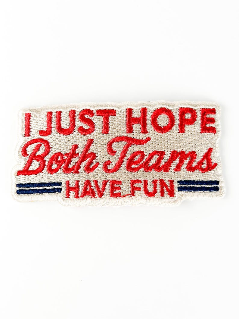 K&ouml;nnte beinhalten: Gestickter Aufn&auml;her mit dem Text "I JUST HOPE Both Teams HAVE FUN" in roter Schrift. Der Aufn&auml;her hat einen beigefarbenen Hintergrund mit zwei blauen Streifen am unteren Rand. Der Aufn&auml;her ist rechteckig.
