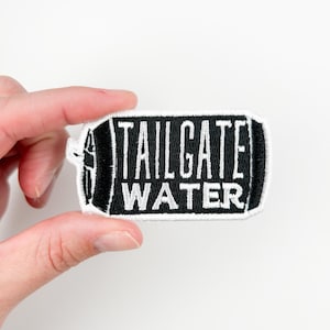 Pode incluir: Patch bordado preto e branco em forma de lata com o texto "TAILGATE WATER" em branco.