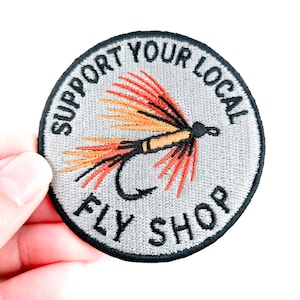 May include: Round embroidered patch with the text "SUPPORT YOUR LOCAL FLY SHOP" in black on a gray background. The patch features a fly fishing lure in orange, yellow, and black.