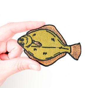 Könnte beinhalten: Ein gestickter Aufnäher mit einem Plattfisch, in brauner und grüner Farbe mit orangefarbenem Umriss.