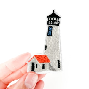 May include: Embroidered patch featuring a lighthouse with a red roof and a tall, gray tower. The lighthouse has a black top and a blue window. The patch is held against a white background.