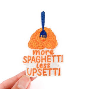 Może przedstawiać: Haftowana naszywka z niebieskim widelcem w pomarańczowym spaghetti. Naszywka zawiera napis "more spaghetti less upsetti" w pomarańczowych literach na białym tle. Naszywka jest trzymana w dłoni.