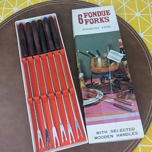 Può includere: Un set di sei forchette per fondue con punte in acciaio inossidabile e manici in legno. Le forchette sono in una scatola bianca con sfondo rosso. La scatola contiene un'immagine di una pentola per fondue e il testo "6 Fondue Forks Stainless Steel With Selected Wooden Handles".