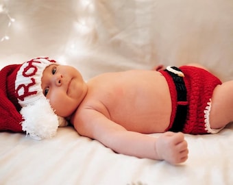 May include: A baby wearing a red and white Santa hat with the word "Santa" embroidered on it, and a red crochet diaper with a black belt. The baby is lying on a white bed with white string lights in the background.