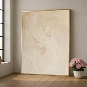 Könnte beinhalten: Großes abstraktes Gemälde in einem goldenen Rahmen. Das Kunstwerk zeigt zwei sich umarmende Figuren, die in neutralen Beige- und Cremetönen dargestellt sind. Ein weißer Keramiktopf mit rosa Blumen steht auf dem Boden vor dem Gemälde.