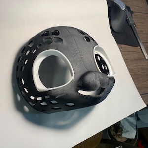 Könnte beinhalten: Eine 3D-gedruckte schwarz-weiße Maske mit einem seehundartigen Design. Die Maske hat große Augenöffnungen, einen lächelnden Mund und zahlreiche Belüftungslöcher. Das Innere der Augen- und Mundöffnungen ist weiß.