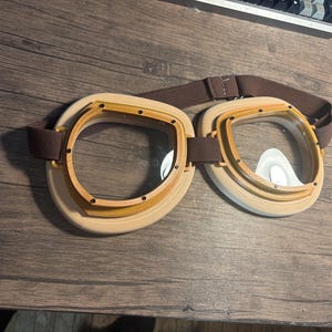 May include: Vintage-style goggles with clear lenses and a brown strap. The goggles have a light brown frame with a darker brown trim. The design is reminiscent of aviator or steampunk aesthetics. The goggles are designed to fit over the eyes.
