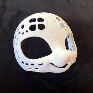 May include: A white, 3D-printed mask with an animal face design. The mask has large eye openings, a prominent nose, and a smiling mouth. It features numerous small, rectangular and circular cutouts across the surface, giving it a cage-like appearance.