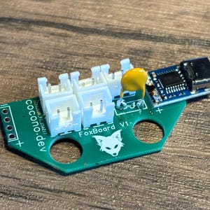May include: A green circuit board with the text "FoxBoard V1" and "cocono.dev" printed on it. The board has a white connector with four pins, a yellow capacitor, and a blue micro-USB connector.