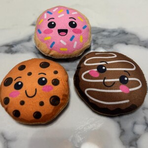 Puede incluir: Tres juguetes de peluche para perros con forma de golosinas. El juguete superior es una rosquilla rosa con chispas y una cara sonriente. Debajo hay una galleta naranja con chispas de chocolate y una cara sonriente. El juguete inferior derecho es una galleta de chocolate con glaseado blanco y una cara sonriente.