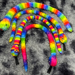 May include: Four colorful plush toy snakes in rainbow hues. The snakes have striped bodies and detailed heads with eyes and black tongues. The plush toys are arranged on a gray, textured surface.