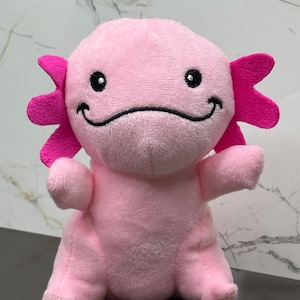 May include: Pink plush axolotl toy with black eyes and a smiling mouth. The axolotl has pink frilly gills and is sitting on a white surface.