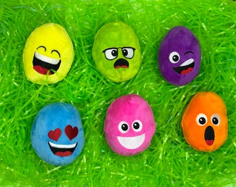 May include: A collection of plush Easter egg toys with various emoji faces. The eggs are yellow, green, purple, blue, pink, and orange, and rest on a bed of green Easter grass.