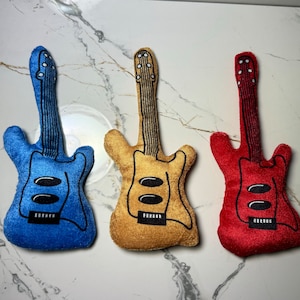 Puede incluir: Tres guitarras eléctricas de peluche en azul, dorado y rojo. Las guitarras tienen detalles bordados en negro y están sobre una superficie de mármol blanco.