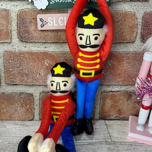 Op de afbeelding: Twee pluche notenkrakerpoppen, één staat met opgeheven armen en de ander zit met gekruiste benen. Beide dragen rood-blauwe uniformen met gouden accenten en hebben zwarte snorren en gele sterren op hun hoeden.