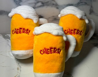 May include: Three plush beer mugs with yellow bodies, white foam tops, and white handles. The word "CHEERS!" is embroidered in red on the front of each mug. The mugs are soft and appear to be made of fabric.