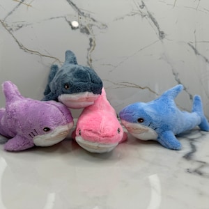 Puede incluir: Cuatro juguetes de peluche con forma de tiburón en diferentes colores: morado, rosa, azul y gris. Los tiburones están todos mirando hacia adelante con la boca abierta.
