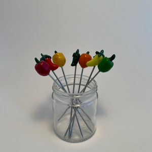 Op de afbeelding: Een glazen pot gevuld met acht metalen spiesjes met kleurrijke paprika- en chilipepervormige toppen. De toppen zijn rood, oranje, geel en groen.