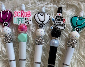 Könnte beinhalten: Vier weiße und schwarze Stifte mit bunten Perlen und Anhängern. Die Anhänger sind herzförmig mit einem Stethoskop und haben den Text "Scrub Life", "Keep Calm I'm a Nurse" und ein türkisfarbenes Herz mit einem Stethoskop.
