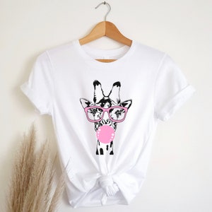 May include: White t-shirt with a black and white illustration of a giraffe wearing pink glasses and blowing a pink bubble.