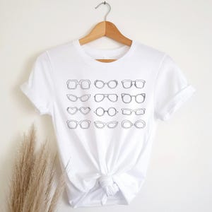 May include: White t-shirt with a black line drawing of various styles of eyeglasses.