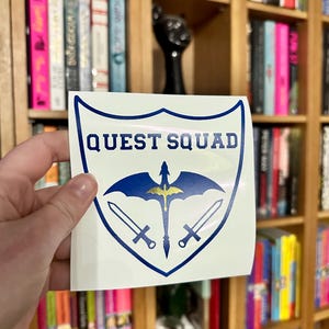 May include: A white vinyl sticker with a blue shield design. The shield features a gold dragon with wings spread and two swords crossed behind it. The text "QUEST SQUAD" is printed in blue letters above the dragon.