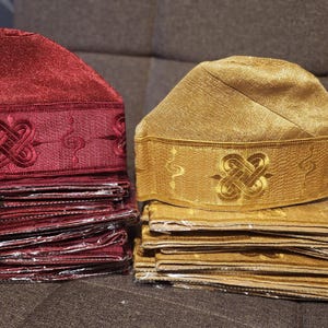 May include: Two embroidered kufi hats, one burgundy and one gold, each atop a stack of matching fabric. The hats feature a decorative band with an intricate knot design. The fabric stacks appear to be the same size as the hats.