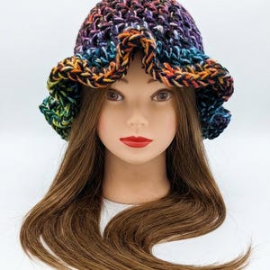Peut inclure: Un chapeau bob au crochet présentant un motif de fil panaché avec des couleurs mélangées de violet, bleu, vert, orange et noir. Le chapeau possède un bord large et ondulé ainsi qu'une construction en maille épaisse posée sur une tête de mannequin.