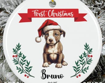 May include: A round white ceramic ornament featuring a brown and white puppy wearing a red Santa hat. Above the puppy is a red banner with the text First Christmas. Below the puppy is the name Bruno and the year 2025, flanked by green holly.