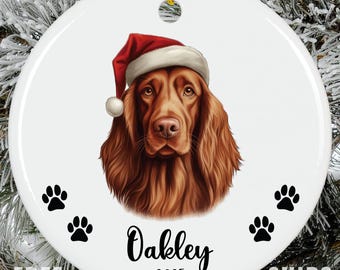 May include: A round white ceramic ornament featuring a detailed illustration of an Irish Setter dog wearing a red and white Santa hat. The name Oakley and the year 2025 are printed in black script below the dog, flanked by four black paw prints.