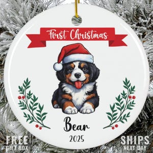 May include: A white ceramic Christmas ornament featuring a Bernese Mountain Dog puppy wearing a Santa hat. The ornament has the text "First Christmas" on a red banner, the name "Bear", and the year "2025". Decorative sprigs and berries frame the puppy.