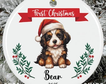 May include: A round white ceramic ornament featuring a Bernese Mountain Dog puppy wearing a red Santa hat. Above the puppy is a red banner with the text First Christmas. Below the puppy is the name Bear and the year 2025, flanked by holly.