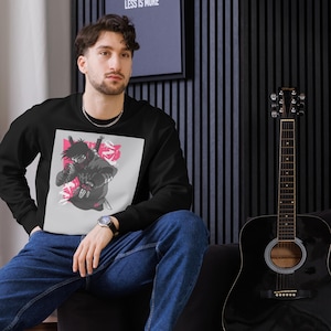 Puede incluir: Sudadera negra de cuello redondo con un gráfico de ninja y texto japonés sobre un fondo blanco y rosa. Una persona lleva la sudadera. A la derecha hay una guitarra negra.