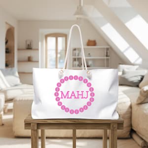 May include: White tote bag with a pink circular design and the letters "MAHJ" in the center. The bag has white rope handles and is sitting on a wooden table. The design is surrounded by pink circles.